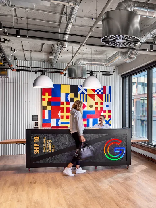 Une personne passe devant la réception d'un immeuble, derrière laquelle on voit une œuvre d'art colorée en forme de drapeau. À droite, de grands néons rouge et orange forment les lettres des mots "Google Montréal".
