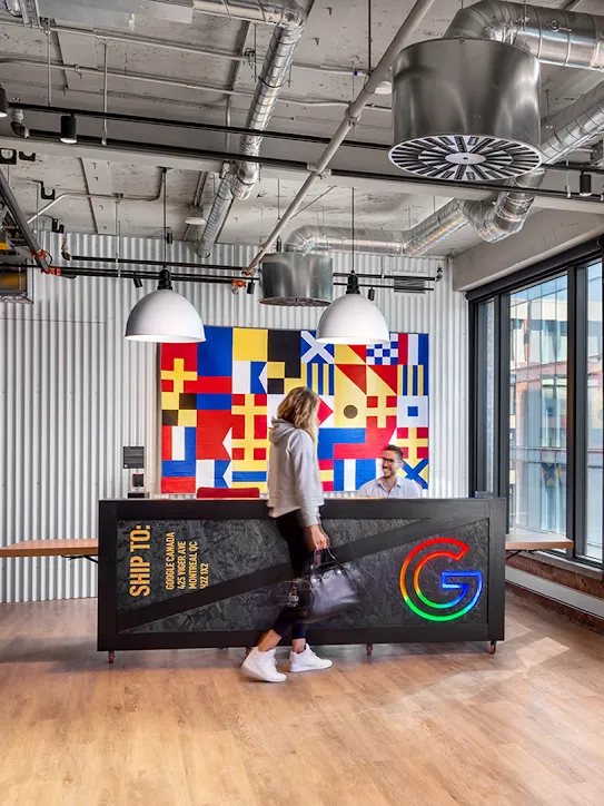 Een persoon loopt langs een balie in een kantoorlobby met een kleurrijk kunstwerk dat op een vlag lijkt. Rechts staan de woorden 'Google Montréal' in grote rode en oranje neonletters.