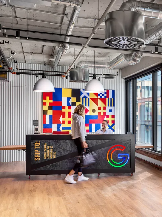 Ofis lobisinde renkli bayrak benzeri bir sanat eserinin bulunduğu resepsiyonun önünden geçen bir kişi. Sağ tarafta büyük kırmızı ve turuncu neon harflerle "Google Montréal" yazıyor.