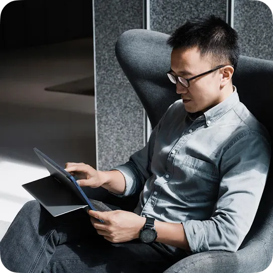 A man in a grey button-down shirt and glasses sits comfortably in a high-backed lounge chair, using the touchscreen of a 2-in-1 laptop resting on his lap.