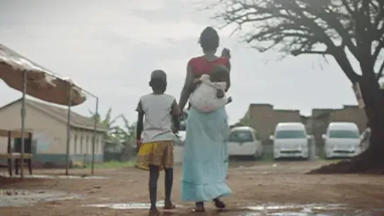 A woman carries her baby on her back and holds her daughter’s hands as they walk away along a dirty road.