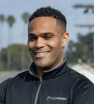 A smiling man wearing a black quarter-zip jacket with the Edvisorly logo on the chest, with palm trees blurred in the background.