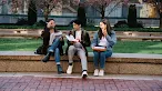 Three young students sit together on a low stone wall in a campus setting, laughing and reviewing papers and notebooks.