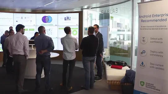 A group of people standing in a "FabLab" innovation center, viewing a large digital wall displaying various data visualizations and charts.