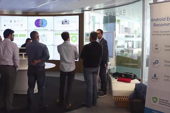 A group of people standing in a "FabLab" innovation center, viewing a large digital wall displaying various data visualizations and charts.