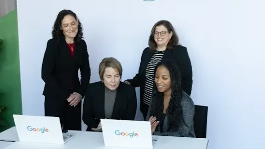 Four women gathered around a white desk with Google-branded laptops during the launch of an AI training initiative. In the background, a white banner displays the Google logo, the Massachusetts state seal, and the Massachusetts AI Hub logo. Two women are seated looking at a laptop screen while the others stand behind them, smiling.