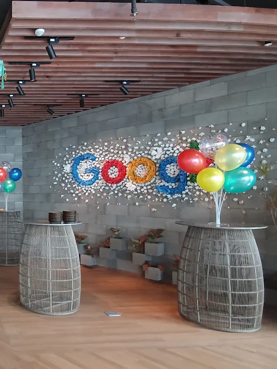 Un espacio de oficina de Google decorado para una celebración. Grupos de globos coloridos se encuentran sobre mesas de estructura metálica y grandes sombrillas de seda cuelgan del techo.