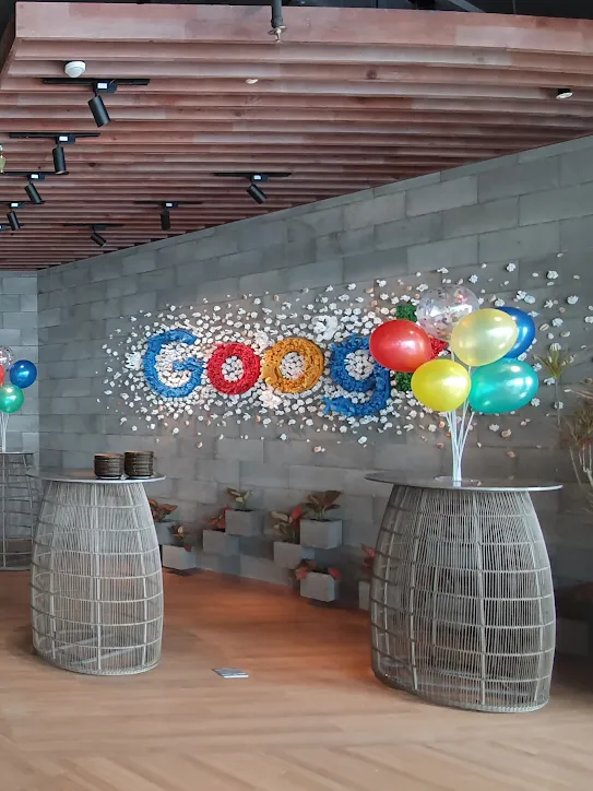A Google office space decorated for a celebration. Groups of colorful balloons sit on wireframe tables and large, silk umbrellas hang from the ceiling.