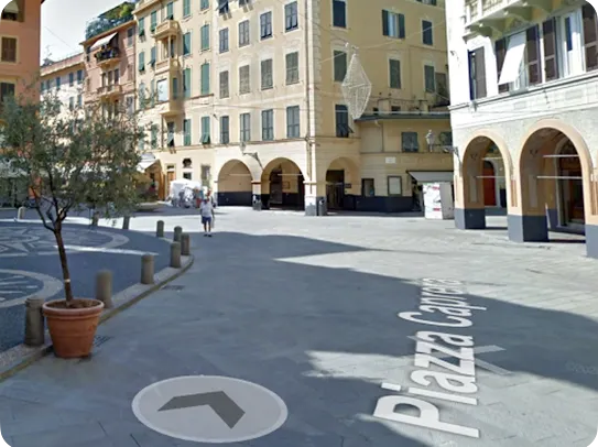 A Google Street View captures an outdoor paved public square. White text reading "Piazza Caprera" and a circular gray navigation arrow are projected onto the gray stone road. Multi-story, peach-colored Italian buildings with green shutters and arched ground-floor porticos surround the open space. A small potted olive tree sits in a terracotta pot near stone bollards in the foreground.