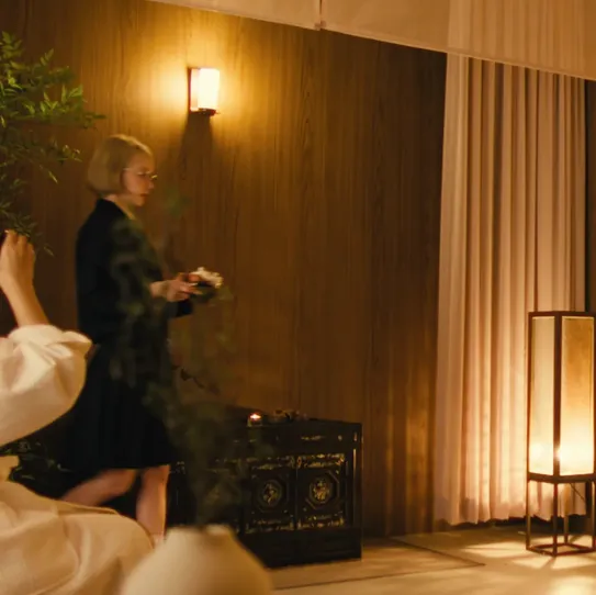 A person in a bathrobe uses their Samsung Galaxy smartphone to ask Gemini to "Play an upbeat song on Spotify." The phone immediately plays "BOOMPALA" by the K-pop group LE SSERAFIM. The music transforms the room into a lively dance party, with the woman now joined by the members of LE SSERAFIM and a group of friends, all dancing energetically to the music.