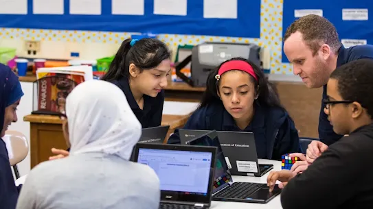 A diverse group of middle school students collaborating on Chromebook laptops at a round table in a classroom. On the right, a male teacher leans in to guide a student wearing glasses, who is typing on his laptop near a Rubik's Cube. In the center, two female students focus intently on a screen together, while two other students on the left, including one wearing a white hijab, participate in the group work. The background shows colorful classroom bulletin boards.