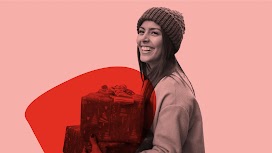 A smiling woman wearing a sweater and a ribbed knit beanie carries two holiday gifts.