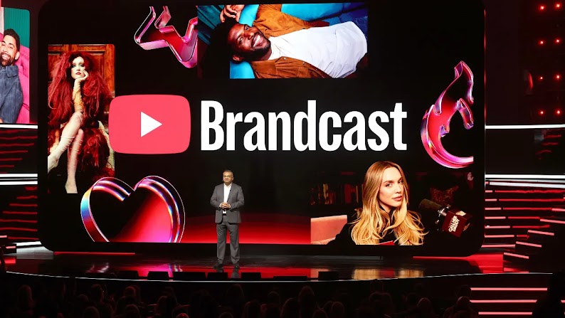 A man with brown skin and short dark hair stands on a stage in front of a large digital screen, showing the Brandcast name and YouTube logo surrounded by images of popular artists and YouTube creators.