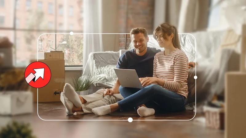 A smiling couple sits on the floor of their new home, surrounded by moving boxes. They are looking at a laptop together. The scene is framed by a white outline with circular handles. A red icon with an upward arrow is in the bottom left.
