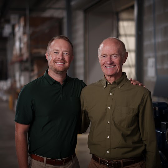 Father and son Norm and Andrew Boyd stand in the Blue Label Packaging warehouse with labeling machinery behind them.