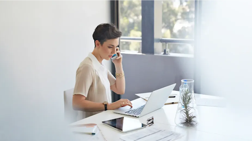 A professional at her desk multitasking on the phone in front of laptop.
