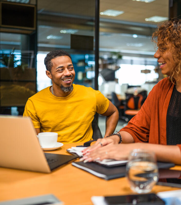Two men and a woman are in a meeting room. They are sitting around a table in front of a laptop, tablet and notebooks, talking to each other and smiling.