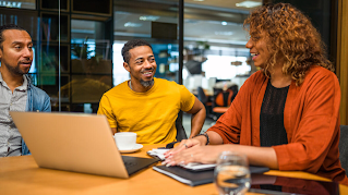 Two men and a woman are in a meeting room. They are sitting around a table in front of a laptop, tablet and notebooks, talking to each other and smiling.