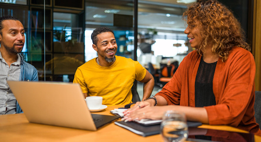 Two men and a woman are in a meeting room. They are sitting around a table in front of a laptop, tablet and notebooks, talking to each other and smiling.