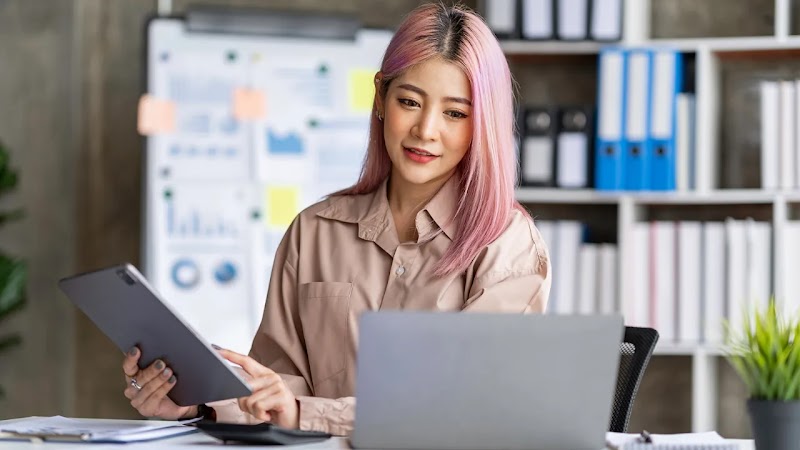 A professional woman with pink hair sits at a desk, looking at a laptop screen while holding a tablet in her other hand. She is in a modern office setting with a whiteboard featuring charts and shelves full of binders in the background.