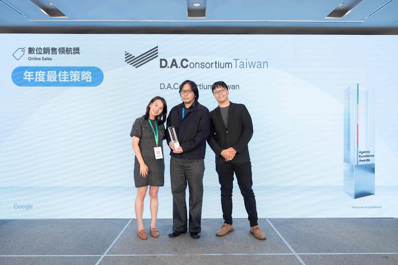 三位業界人士於 Google 獲獎典禮舞台領取獎項。背景展示「數位銷售領航獎」與「年度最佳策略」藍色標籤，中央為「D.A.C Consortium Taiwan」黑色標誌與文字。左上角有「數位銷售領航獎」與「Online Sales」標籤，Google 標誌在左下方，右側為獎項標牌。獲獎者穿著深灰色連身裙、深藍色西装與黑色商務服飾，配戴證件掛牌。台灣迪艾思(HIMT)為國際高端珠寶品牌 Mikimoto 導入雙軌成長策略，透過 Google AI 驅動的數位銷售優化，同步提升實際收益與降低進站成本，在數位場域創造新的競爭優勢。
