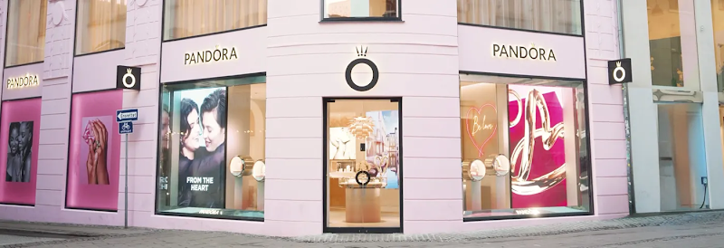 Exterior of a pink Pandora jewelry store on a cobblestone street. The storefront features glowing PANDORA logos, large display windows with "FROM THE HEART" and "Be love" signage, and the brand's crown O emblem above the entrance.
