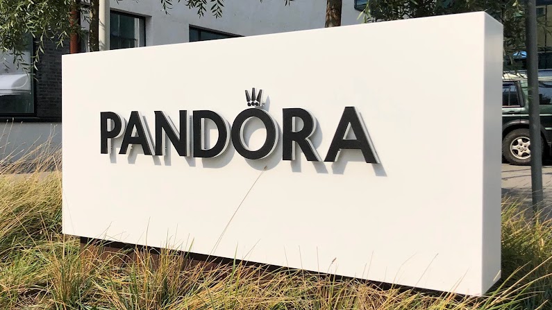 A large white outdoor sign displaying the black 3D "PANDORA" logo topped with its signature crown symbol, set among ornamental grasses outside a building.