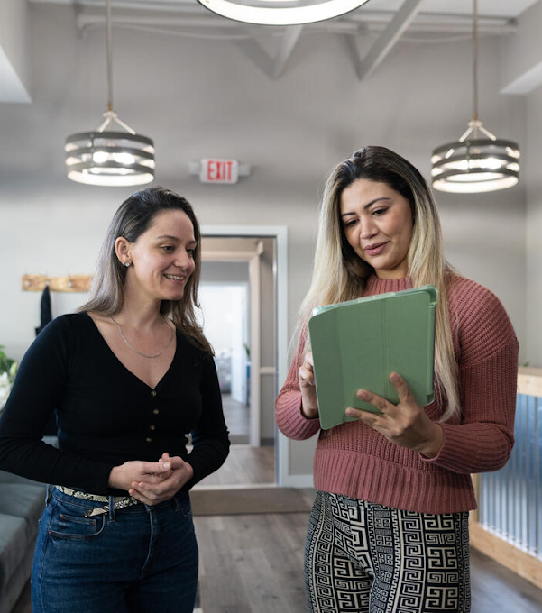 Two women are standing next to each other. The woman on the right is holding a tablet and discussing its content with the other woman. 