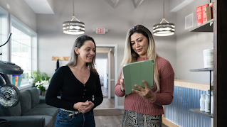 Two women are standing next to each other. The woman on the right is holding a tablet and discussing its content with the other woman. 