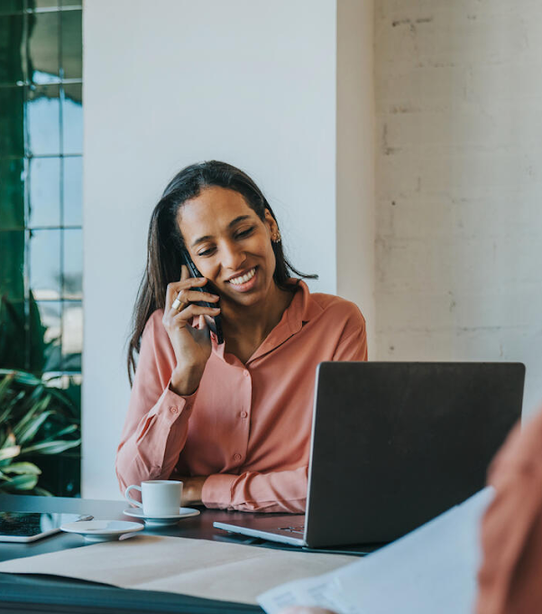 A woman is talking on the phone, smiling. A laptop, coffee cup and tablet are on the table in front of her, and a man photographed from the back is on the opposite side of the table.