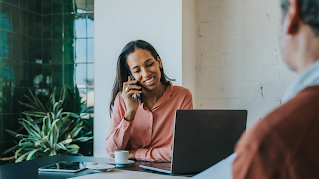 A woman is talking on the phone, smiling. A laptop, coffee cup and tablet are on the table in front of her, and a man photographed from the back is on the opposite side of the table.