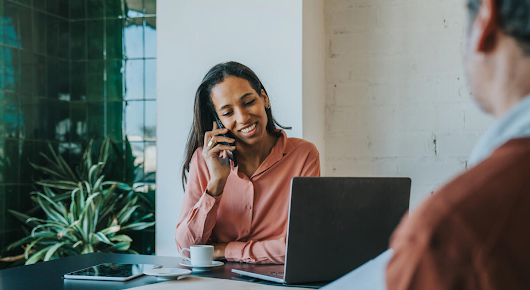 A woman is talking on the phone, smiling. A laptop, coffee cup and tablet are on the table in front of her, and a man photographed from the back is on the opposite side of the table.