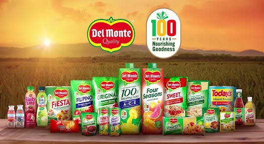 A collection of various Del Monte food and beverage products arranged on a wooden table against a sunrise field background, with logos celebrating Del Monte's 100th anniversary.