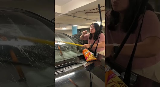 The video thumbnail shows a young woman in a pink top cleaning the windshield of a black car in an underground parking garage. She is holding a long, yellow squeegee or brush and appears to be concentrating while cleaning. A bag of Nova multigrain snacks is resting on the car's hood near the windshield wiper.