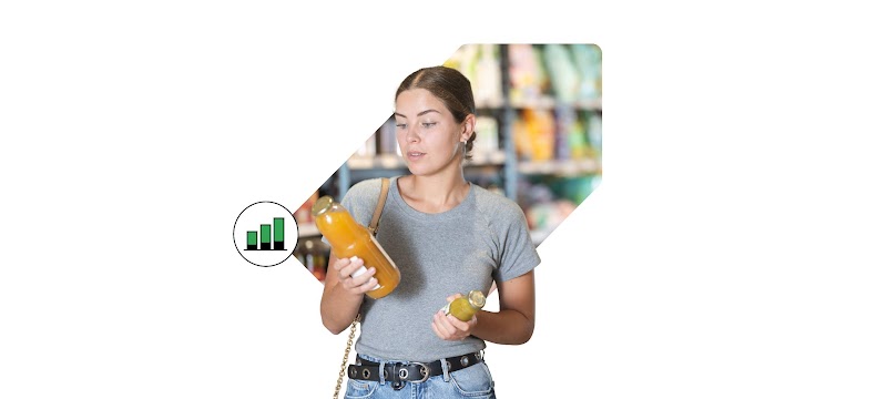 Una mujer con el pelo rubio recogido, con camiseta gris y vaqueros, en un supermercado compara dos botellas de zumo junto a un icono de un gráfico de barras que representa el análisis de datos de consumo.