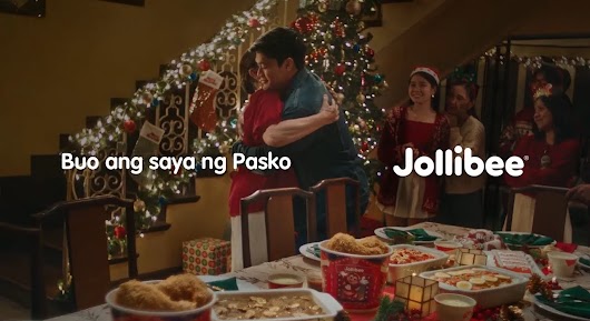 A Jollibee Christmas advertisement showing a family gathered around a holiday feast, with a couple hugging in front of a Christmas tree. Text on the screen reads 'Buo ang saya ng Pasko' alongside the Jollibee logo