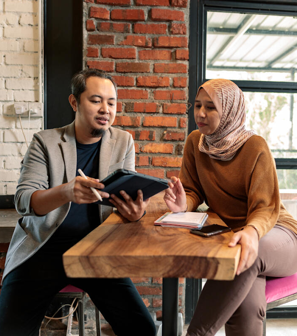 A man and a woman are sitting on opposite sides of a table. The man is holding a tablet and digital pen, and the woman has a phone and notebook in front of her.