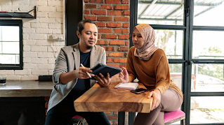A man and a woman are sitting on opposite sides of a table. The man is holding a tablet and digital pen, and the woman has a phone and notebook in front of her.
