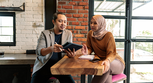 A man and a woman are sitting on opposite sides of a table. The man is holding a tablet and digital pen, and the woman has a phone and notebook in front of her.