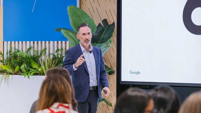 Sean Downey, president, Americas and Global Partners at Google, speaks during CMO AI Lab in New York City. A planter with palm plants and a large screen are behind Downey, who has light skin, graying dark hair, and wears a sport coat with slacks.
