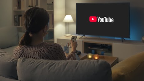 Donna seduta sul divano di spalle mentre tiene in mano un telecomando puntato verso la TV, sullo schermo appare il logo di YouTube in una stanza illuminata da luci soffuse.