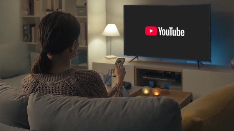 Donna seduta sul divano di spalle mentre tiene in mano un telecomando puntato verso la TV, sullo schermo appare il logo di YouTube in una stanza illuminata da luci soffuse.