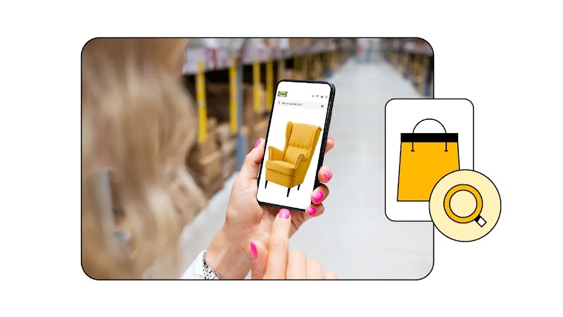 A woman browses the IKEA website on her smartphone, inside an IKEA store. The screen displays the image of a yellow sofa. An icon of a yellow shopping bag with a magnifying glass overlay on the right.