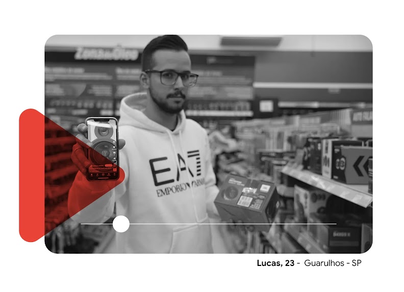Foto de Lucas, em uma loja de eletrodomésticos, mostrando seu celular com vídeos de resenhas de produtos. Sobre a foto em p&b tem um ícone de play vermelho.