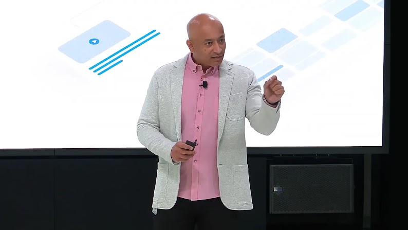 Tarun Rantham, global director, AI and Cloud for marketers, stands in front of a digital screen, giving a talk. Rantham has medium skin, no hair, and wears a gray sport coat over a pink button-front shirt with black pants.