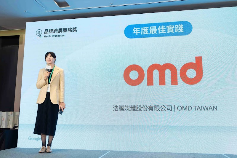 浩騰媒體代表於 Google 獲獎典禮舞台上發表。背景展示「品牌跨屏策略獎」與「年度最佳實踐」藍色標籤，紅色 OMD 字體與公司全名「浩騰媒體股份有限公司 OMD TAIWAN」。左上角有「品牌跨屏策略獎」與「Media Unification」標誌，Google 標誌在左下方。講者穿著淡黃色西裝、深色長裙，展示 YouTube 跨屏策略核心，透過全格式整合與 AI 驅動最佳化，達成短期爆發與長期成長同步提升的跨屏影音成效。