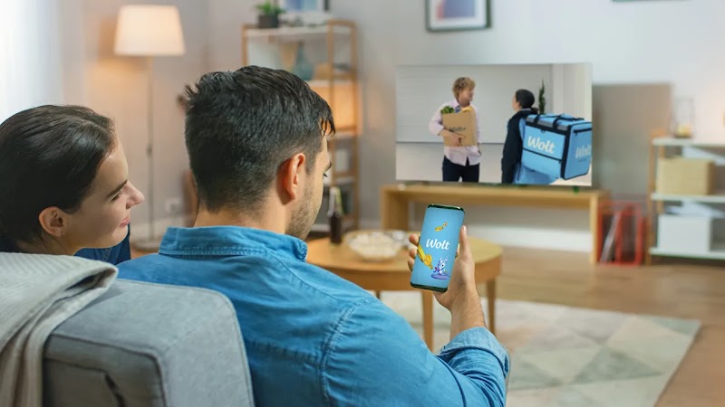 A couple on a sofa watches a TV advertisement for Wolt delivery. The man holds a smartphone displaying the Wolt app, subtly indicating the ad's direct influence on their engagement with the service.