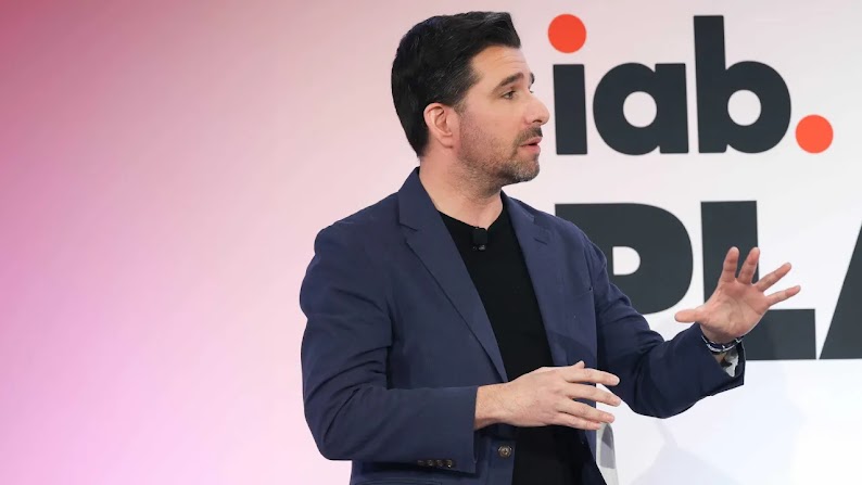 Jack Koch, SVP di IAB di ricerca e statistica, parla sul palco a una conferenza. Koch è seduto su uno sgabello alto e gesticola con le mani mentre parla. Ha la pelle chiara, i capelli corti scuri e indossa una giacca nera, una camicia nera senza colletto