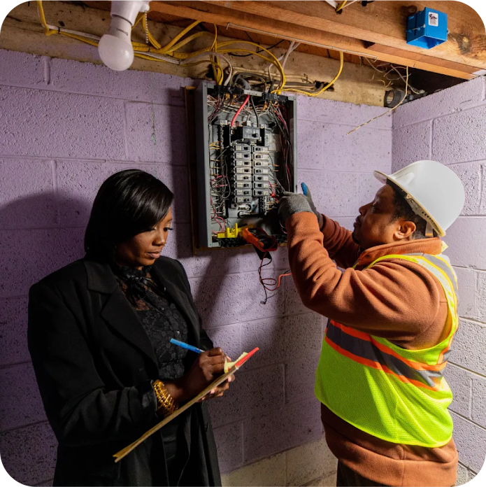 A woman writes on a clipboard while an electrician wearing a safety vest and hardhat works on a breaker box 