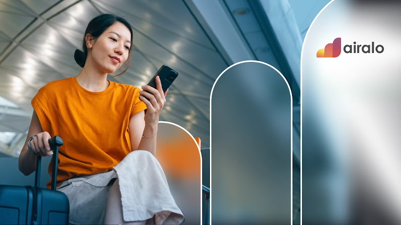 A woman with light skin and dark hair uses her mobile phone while sitting with her suitcase in an airport waiting area. The Airlo wordmark appears to her right.